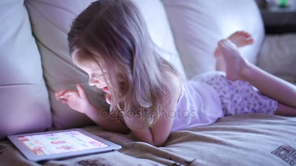 Niña linda sentada en el sofá y jugando con la tableta — Vídeos de Stock