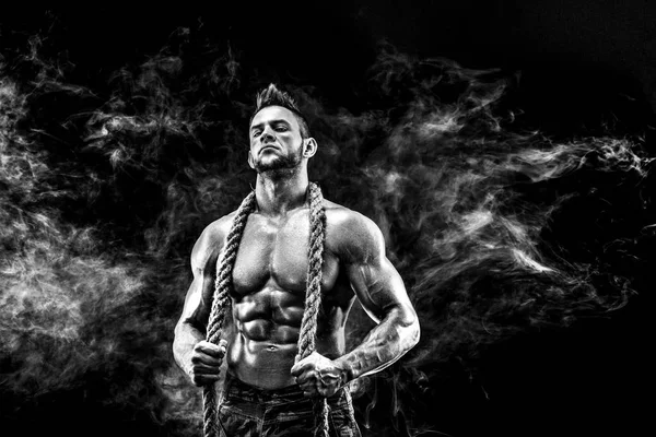 Strong athletic man with naked body in military pants and rope on neck dark with smoke background — Stock Photo, Image