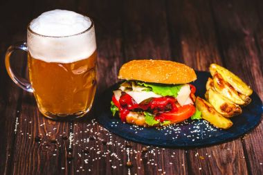 Ev yapımı lezzetli, ağız sulandıran sığır eti burger marul ve patates, taş kesme tahtası üzerinde bardak bira servis. Koyu ahşap arka plan