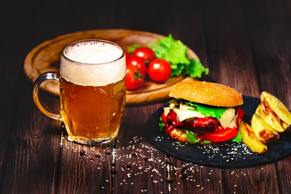 Hamburguesa casera deliciosa y deliciosa con lechuga y patata, vaso de cerveza servido en una tabla de cortar piedra. Fondo de madera oscura — Foto de Stock