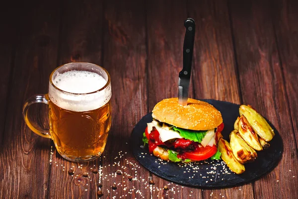 Home made mouth-watering, delicious beef burger with lettuce and potato, glass of beer served on stone cutting board. Dark wooden background — Stock Photo, Image