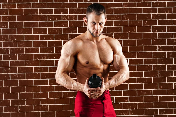 Ung muskulös man visar sin perfekta kropp. en man som dricker från en cocktail shaker med proteinet mot tegelvägg — Stockfoto