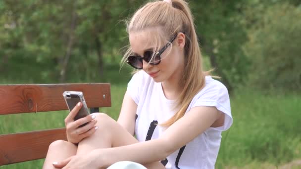 Meisje praten op de slimme telefoon zit in een park op houten bankje met groene boom blad, natuur scène — Stockvideo