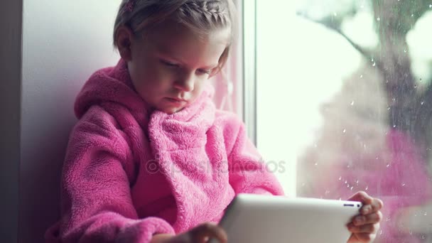 Linda niña en albornoz rosa usando la tableta PC mientras está sentado en el alféizar de la ventana . — Vídeo de stock