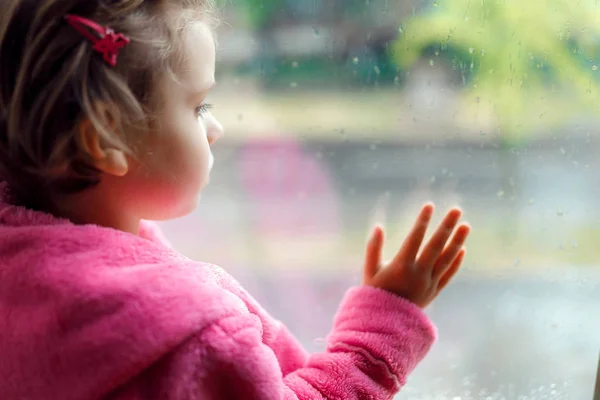 Zbliżenie na Little cute girl w różowy szlafrok patrzy niestety z okna. Siedząc na parapecie. — Zdjęcie stockowe