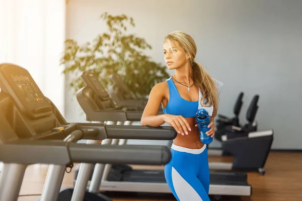 Hermosa mujer rubia atlética está bebiendo agua en una cinta de correr en el gimnasio . —  Fotos de Stock