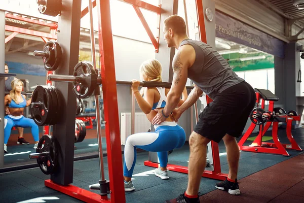 Sporty girl doing squat exercises with assistance of her personal trainer at gym. — Stock Photo, Image