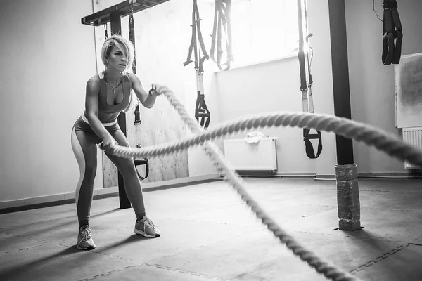 Woman with battle ropes exercise in the fitness gym. Young female wearing sportswear.