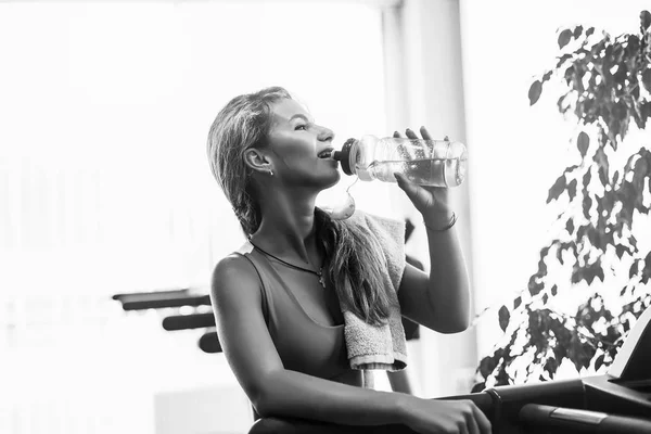 Belle femme blonde athlétique boit de l'eau sur un tapis roulant dans la salle de gym . — Photo