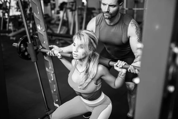 Sporty girl doing squat exercises with assistance of her personal trainer at gym. — Stock Photo, Image