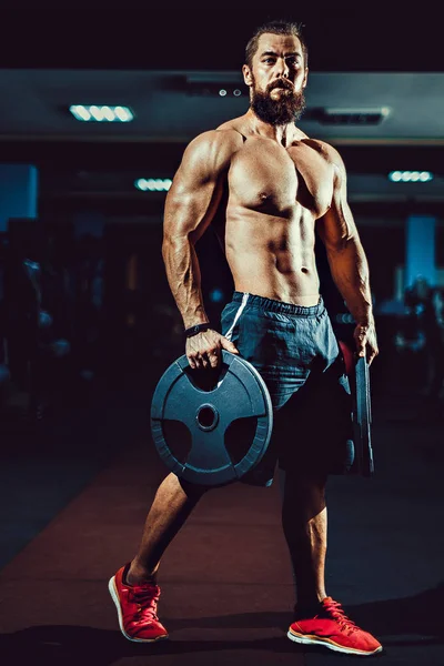 Atleta musculoso fisiculturista homem posando com halteres no ginásio . — Fotografia de Stock