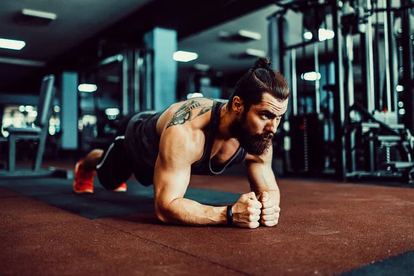 Hombre joven musculoso que usa ropa deportiva y hace posición de tablón mientras hace ejercicio en el suelo en el interior del loft — Foto de Stock