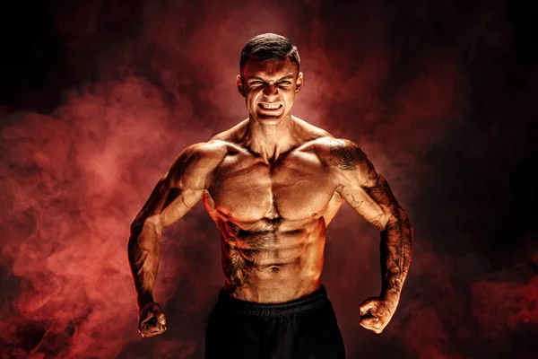 Culturista posando. Fitness hombre musculoso tatuado sobre fondo de humo rojo . — Foto de Stock