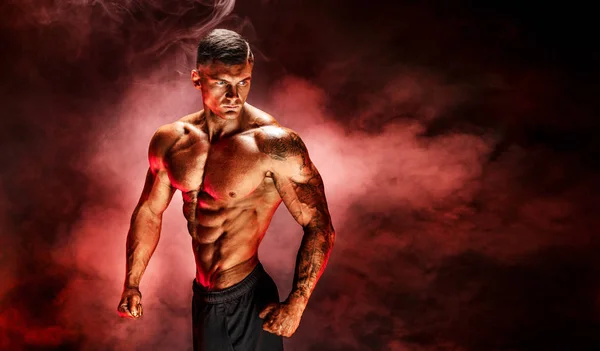 Bodybuilder posing. Fitness tattooed muscled man on red smoke background.