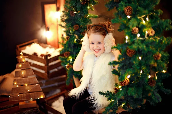 Menina bonito em earmuffs de pele sentado perto da árvore de Natal — Fotografia de Stock