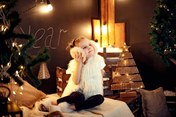 Söt liten flicka i päls öronmuffar sitter nära julgran — Stockfoto