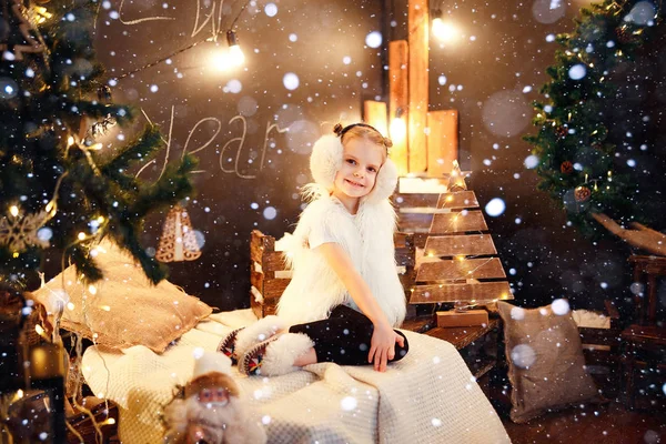 Söt liten flicka i päls öronmuffar sitter nära julgran — Stockfoto