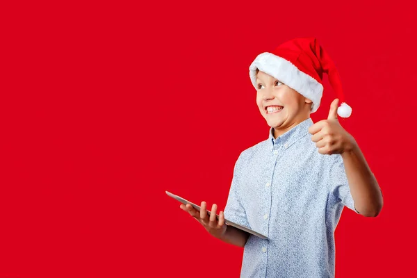 Joven santa boy haciendo el ok pulgares hacia arriba gesto mientras sostiene una tableta —  Fotos de Stock