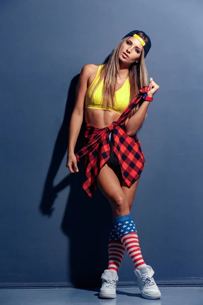 Sexy hermosa modelo de mujer joven en camisa deportiva y sombrero, camisa a cuadros rojo, reloj de pulsera, zapatillas blancas, leggings con bandera de EE.UU., apoyado en la pared texturizada . — Foto de Stock