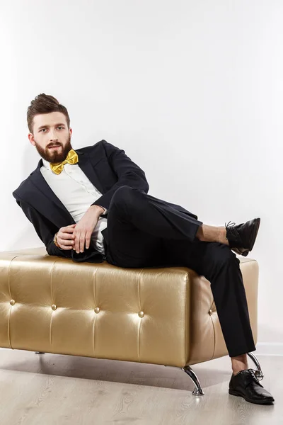 Homem bonito jovem elegante em traje clássico preto. Retrato de moda de estúdio. Sente-se no sofá dourado . — Fotografia de Stock