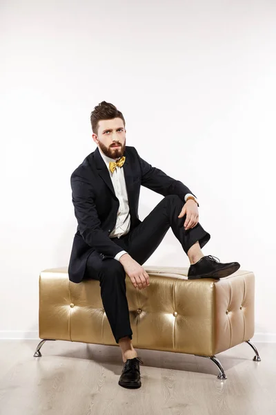 Homem bonito jovem elegante em traje clássico preto. Retrato de moda de estúdio. Sente-se no sofá dourado . — Fotografia de Stock