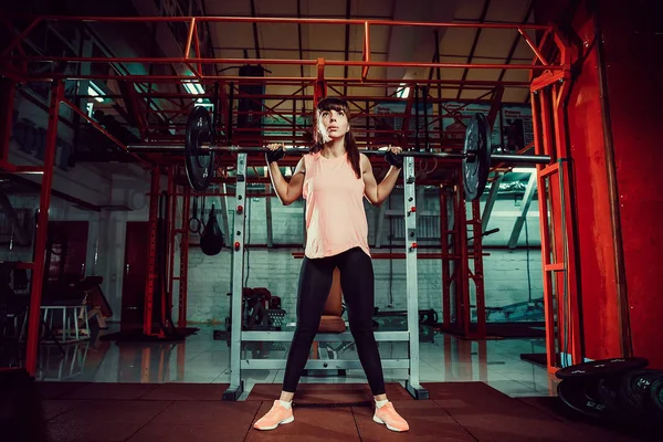 Hinreißende junge Fitness-Frau hockt und macht Kadertraining mit einer Stange hinter dem Hals in der Turnhalle. — Stockfoto