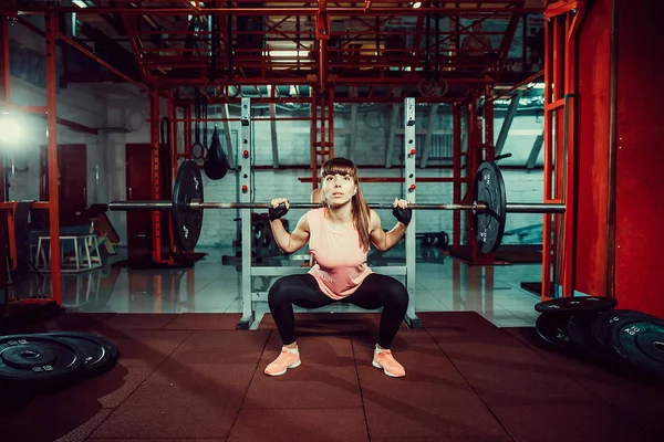 Hinreißende junge Fitness-Frau hockt und macht Kadertraining mit einer Stange hinter dem Hals in der Turnhalle. — Stockfoto