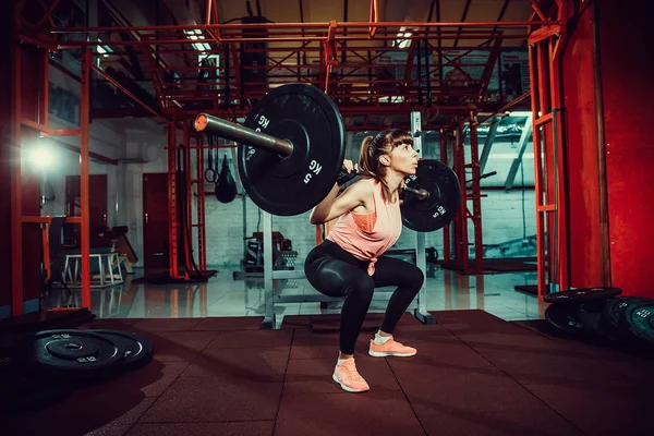 Hinreißende junge Fitness-Frau hockt und macht Kadertraining mit einer Stange hinter dem Hals in der Turnhalle. — Stockfoto