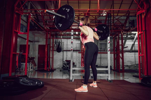 Hermosa joven fitness mujer agacharse y hacer ejercicio de escuadrón con una barra detrás del cuello en el gimnasio . — Foto de Stock