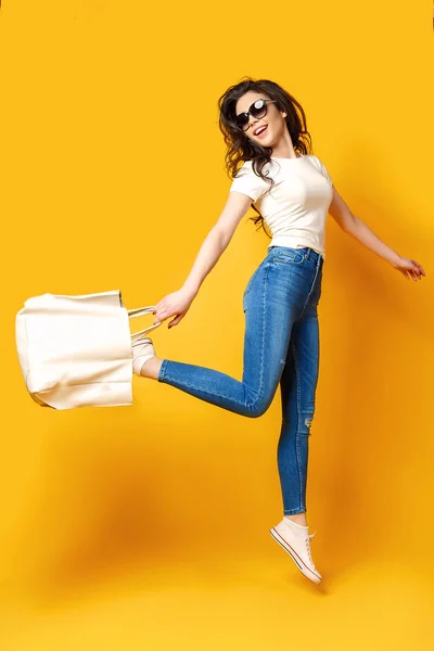 Mulher bonita em óculos de sol, camisa branca, jeans azul pulando com saco no fundo amarelo — Fotografia de Stock