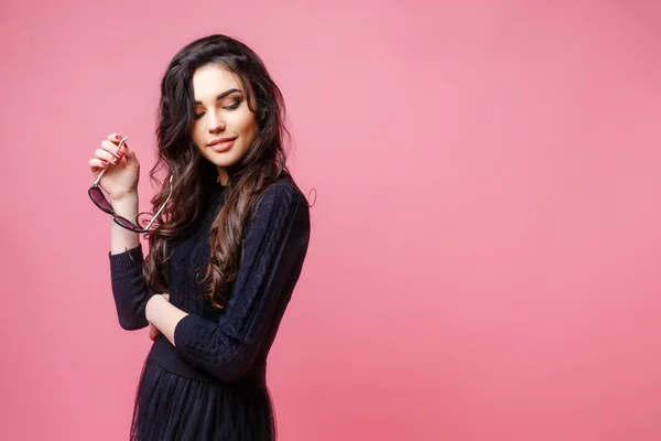 Young pretty sexy woman or girl with cute face and long brunette hair wearing sunglasses and black sweater, posing on pink background — Stock Photo, Image