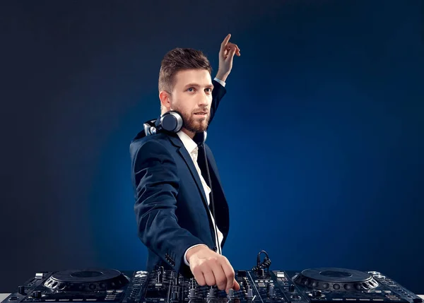 Retrato de close-up de DJ confiante com estilo de cabelo elegante e fones de ouvido no pescoço misturando música no mixer enquanto estava isolado em azul escuro, fundo ciano — Fotografia de Stock