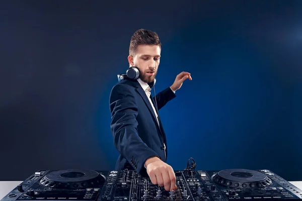 Primer plano retrato de DJ confiado con estilo de pelo elegante y auriculares en el cuello mezclando música en el mezclador, mientras que de pie aislado en azul de color oscuro, fondo cian —  Fotos de Stock