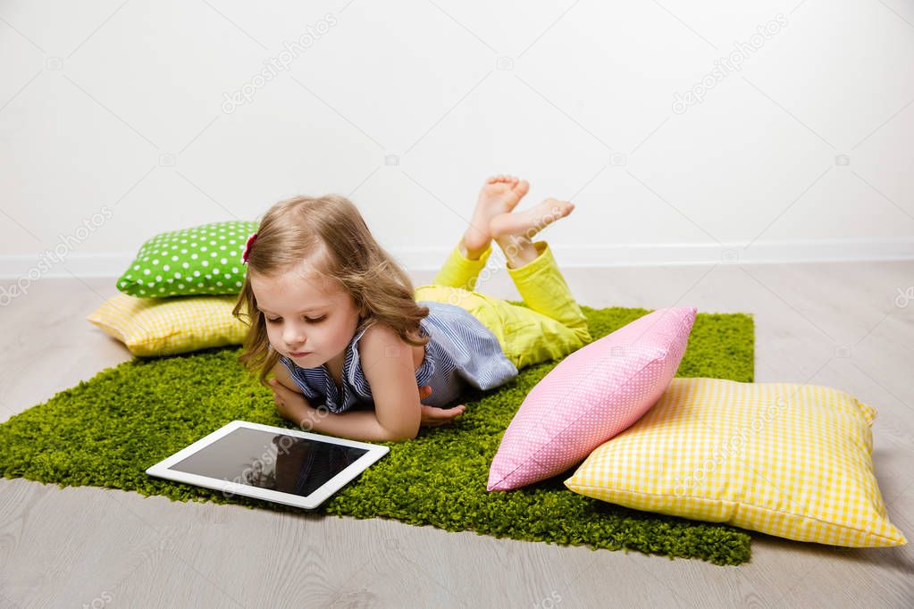 The little girl lies on a rug, watches the tablet.