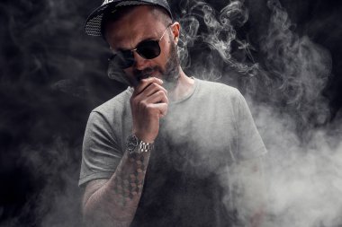 Sakallı erkek gri gömlek, güneş gözlüğü ve beyzbol kap vaping giymiş