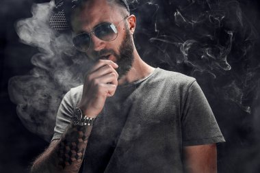 Sakallı erkek gri gömlek, güneş gözlüğü ve beyzbol kap vaping giymiş