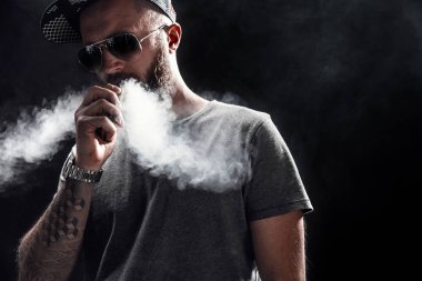 Sakallı erkek gri gömlek, güneş gözlüğü ve beyzbol kap vaping giymiş