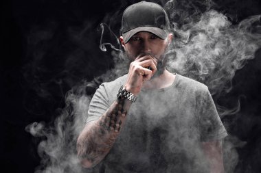 Sakallı erkek gri gömlek, güneş gözlüğü ve beyzbol kap vaping giymiş