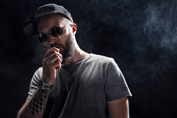 Bearded male dressed in a grey shirt, sunglasses and baseball cap vaping — Stock Photo, Image