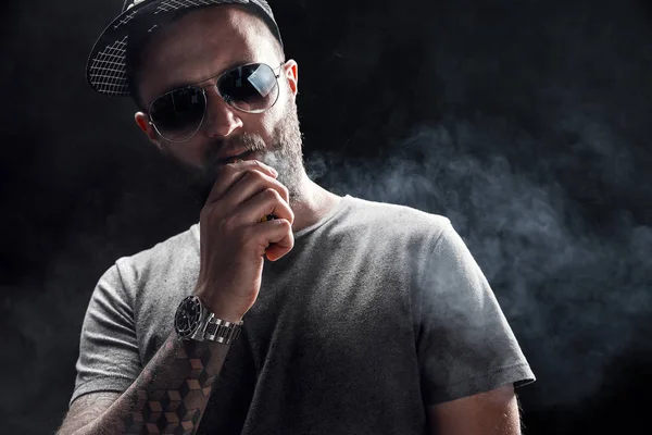 Bearded male dressed in a grey shirt, sunglasses and baseball cap vaping — Stock Photo, Image