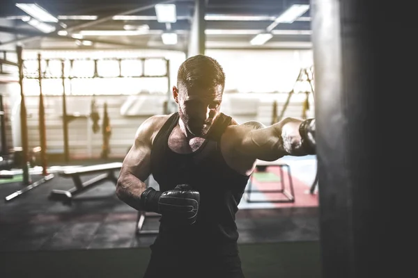 Mężczyzna bokser trening z workiem bokserskim w ciemnej hali sportowej. — Zdjęcie stockowe