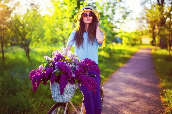 Hermosa joven con bicicleta vintage y flores en el fondo de la ciudad a la luz del sol al aire libre . — Foto de Stock