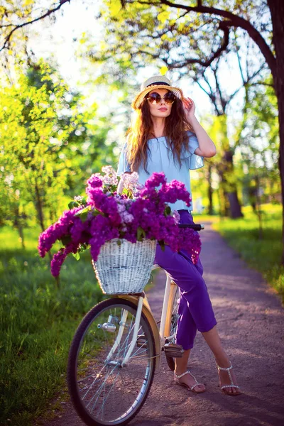 Hermosa joven con bicicleta vintage y flores en el fondo de la ciudad a la luz del sol al aire libre . —  Fotos de Stock