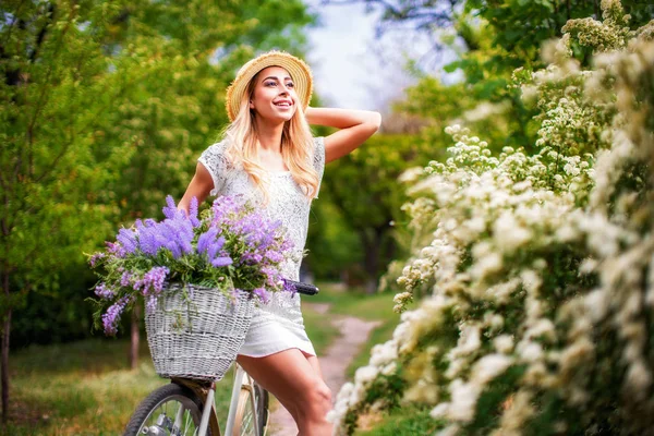 Hermosa joven con bicicleta vintage y flores en el fondo de la ciudad a la luz del sol al aire libre . —  Fotos de Stock