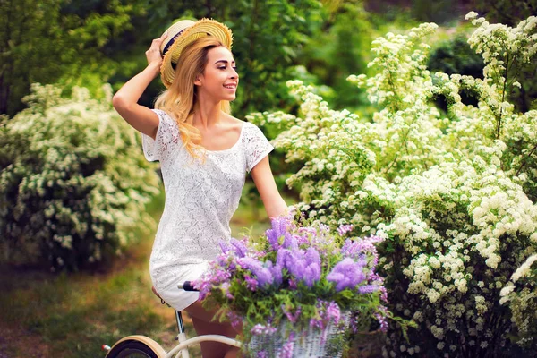 Hermosa joven con bicicleta vintage y flores en el fondo de la ciudad a la luz del sol al aire libre . —  Fotos de Stock