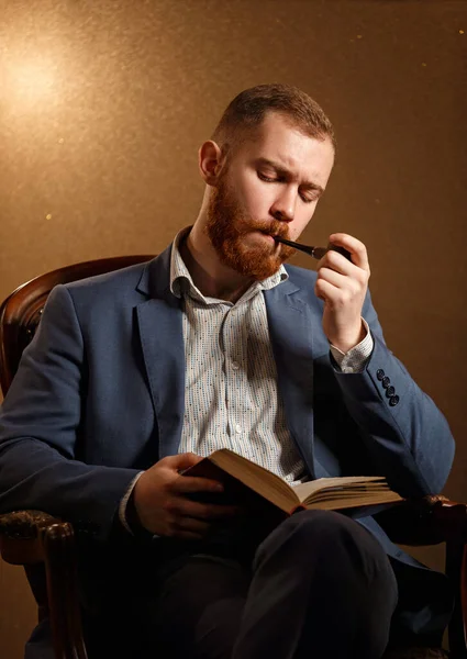 Barbudo Lamber como modelo masculino en traje con bigote y barba fumar pipa y leer libro mientras está sentado en la silla — Foto de Stock