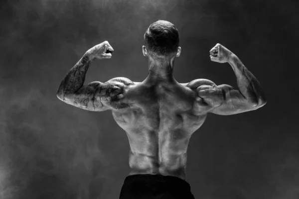 Vista posterior del hombre irreconocible, músculos fuertes posando con los brazos abajo en humo . — Foto de Stock