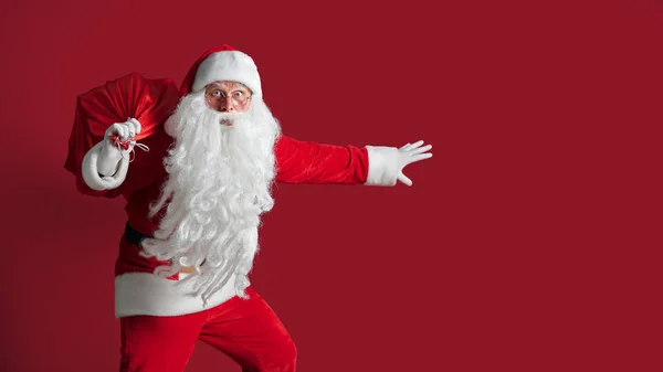 Portret van verrast hurken Santa Claus dragen enorme rode zak met geschenken op rode studio achtergrond. isolaat — Stockfoto