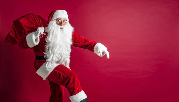 Porträt eines hockenden Weihnachtsmannes, der einen riesigen roten Sack mit Geschenken auf rotem Studiohintergrund trägt. isolieren — Stockfoto