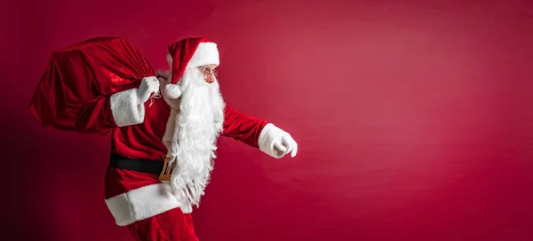 Портрет приседающего Санта-Клауса с огромным красным мешком с подарками на красном фоне студии. Изолировать — стоковое фото